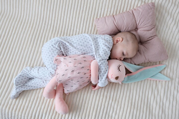 Baby with pink bunny soft toy. A girl of European appearance, wearing a light light jumpsuit, is sleeping in the crib. Sleep mode, care and hygiene.