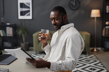 Side view portrait of successful African American entrepreneur holding glass of whiskey while reading documents at workplace in office, copy space
