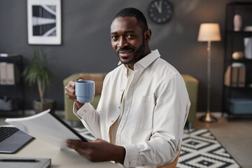 Portrait of smiling African American man looking at camera at home office and holding coffee cup,...