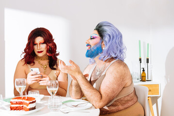 Happy adult man drag queen in colorful wig sitting at table with focused friend using smartphone