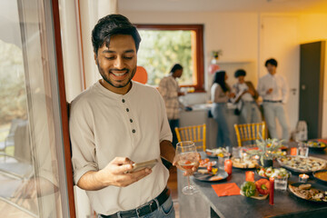 Fototapeta na wymiar Smiling man with glass of wine and phone standing on friends background at home holiday party