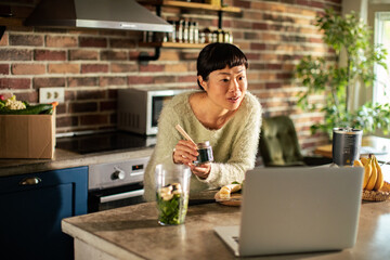 Asian woman making smoothie while working on laptop in kitchen
