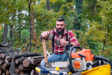 Caucasian man, a lumberjack, takes a moment to rest on a pile of firewood, pouring himself a cup of coffee from a thermos, his tools laid out in front of him.