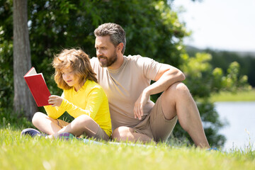 Fototapeta premium Father and son enjoy spending quality time reading books outdoors. Portrait of father and son relax on green lawn in summer park and read book together. Father and his little son reading book in park.