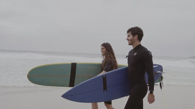 Casal de surfistas caminhando na praia com as pranchas debaixo do braco, conversando. Cinematografico 4k.