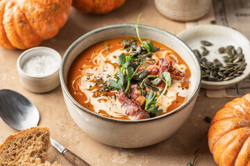 Bowl of pumpkin cream soup decorated with cream, pumpkin seeds, bacon, microgreens and olive oil on brown textured background with ingredients above