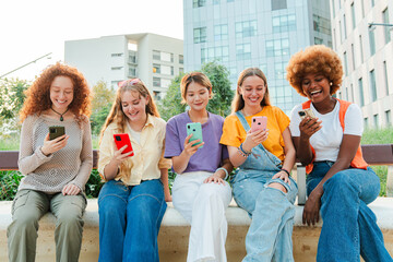 Multiracial group of young female friends enjoying and smiling using their mobile phone app sitting...