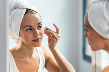 Beautiful young woman removing makeup while taking care of her skin while looking at mirror at home.