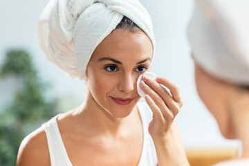 Beautiful young woman removing makeup while taking care of her skin while looking at mirror at home.