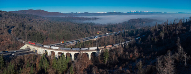 Modern logistics - single train running over the massive viaduct over motorway with trucks driving...