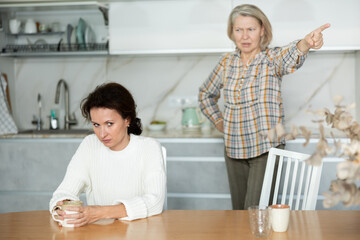 Upset middle-aged woman sitting at the kitchen table with her back to old woman quarreling angrily...