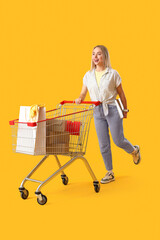 Young woman with laptop and market trolley full of shopping bags on yellow background