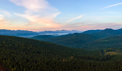 Aerial view of bright foggy morning over dark hills with mountain forest trees at autumn sunrise. Beautiful scenery of wild woodland at dawn