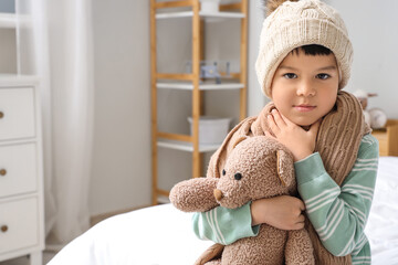 Ill little Asian boy with toy bear suffering from sore throat in bedroom