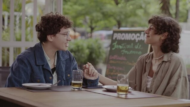 Duas mulheres sentadas &agrave; mesa de um restaurante ao ar livre, conversando e de maos dadas. Cinematico 4k.