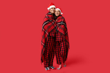 Happy young couple in Christmas pajamas and Santa hats wrapped in warm plaid on red background