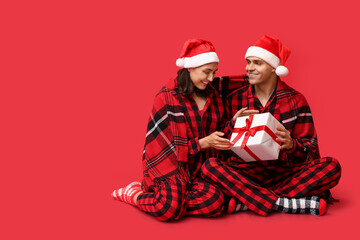 Happy young couple in Christmas pajamas and with gift on red background