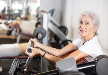 Mature woman pumps up her arm muscles on a special exercise machine in the gym