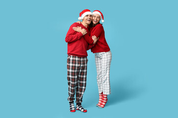 Happy young couple in Christmas pajamas and Santa hats hugging against blue background