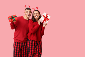 Happy young couple in Christmas pajamas and with gifts on pink background