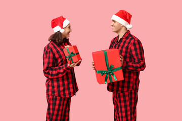 Happy young couple in Christmas pajamas and with gifts on pink background