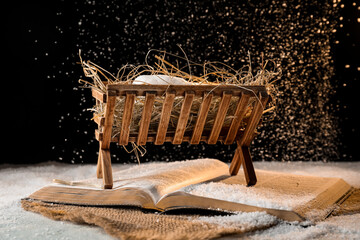 Bible, wooden manger with hay and dummy of baby on snow against black background. Concept of...