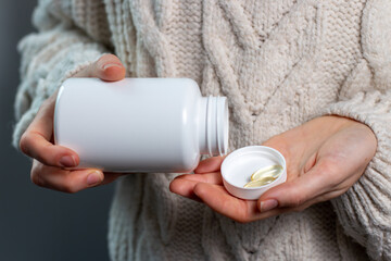 Woman hand holds yellow medication capsules of omega 3, vitamin D, pours from a white bottle into...