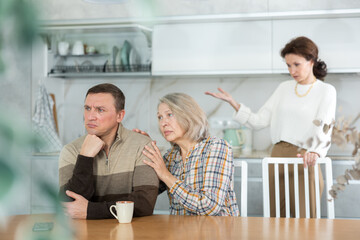 Family members quarreling in the kitchen and old woman trying to calm angry man sitting at the table