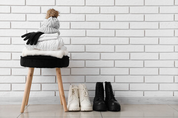 Stack of folded sweaters with hat, gloves on chair and boots near white brick wall - obrazy, fototapety, plakaty