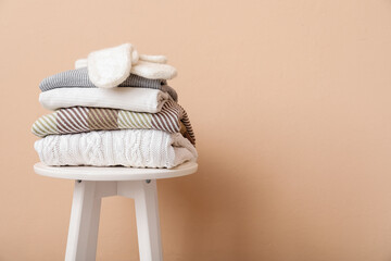 Stack of folded sweaters with gloves on stool against beige background