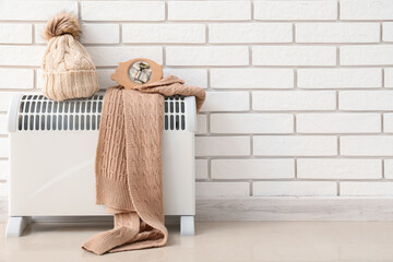 Electric radiator with piggy bank, hat and scarf near white brick wall. Heating saving concept