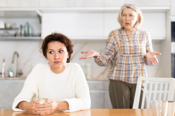 Upset middle-aged woman sitting at the kitchen table with her back to old woman quarreling angrily...