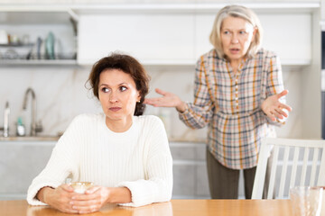 Sad middle-aged woman sitting with a cup in hand and mature female family member brawling to her in...