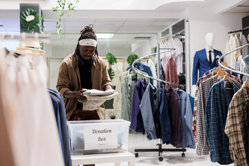 African american man putting clothing in donation box in shopping mall. Fashion boutique customer packing apparel in container for charity organization, giving garment for poor - obrazy, fototapety, plakaty