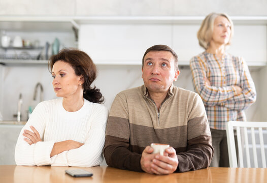 Angry Middle-aged Man And Woman Sitting At Table And Upset Old Woman Standing Behind In The Kitchen