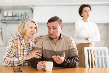 Aggrieved middle-aged man sitting at the kitchen-table after brawling with wife and old woman speaking to him peacefully