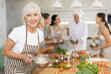 Elderly woman in apron learning to cook at cooking master class - obrazy, fototapety, plakaty