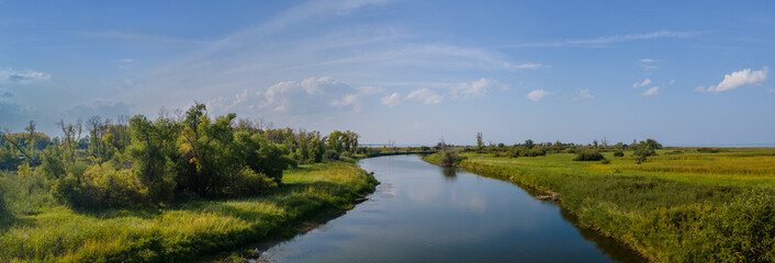 Panoramic view along a quiet curving river that is surrounded by green grass and small shrubs under...