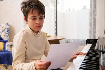 Adorable multi ethnic teenage boy learning musical notes, sitting at piano and performing classical...