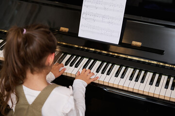Rear view little child girl performing the rhythm of classical music while playing grand piano,...