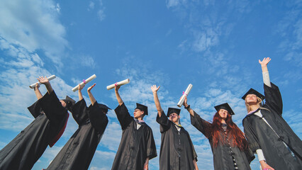 Cheerful graduates pose with raised diplomas on a sunny day.