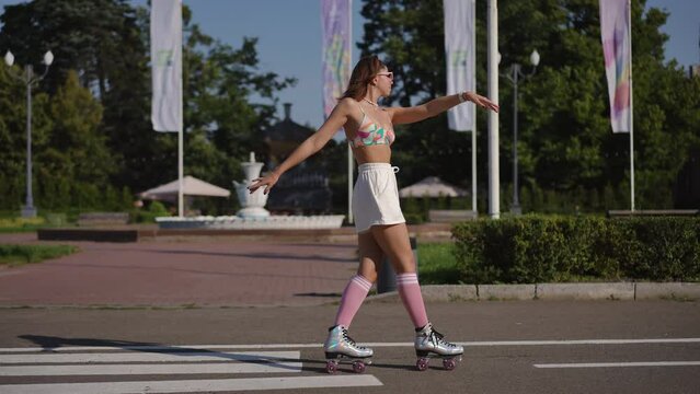 A young woman in glasses dances elegantly on roller skates against the backdrop of a city park on a sunny day outdoors. Slow motion