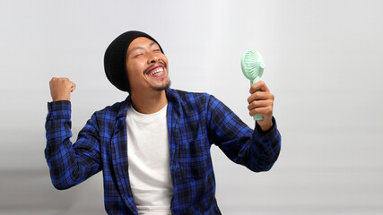 Asian man, dressed in a beanie hat and casual shirt, revels in the airflow and refreshing cool wind from a handheld portable mini fan to beat the summer heat, while standing against a white background