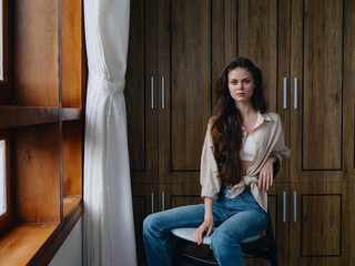 Woman sitting on a chair in home interior near the window, comfort and coziness, lifestyle relaxation on the weekend, pensive look of a beautiful girl.