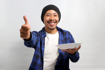 Cheerful young Asian man, dressed in a beanie hat and casual shirt, expresses happiness and...