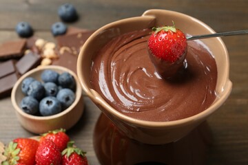 Dipping fresh strawberry in fondue pot with melted chocolate at wooden table, closeup