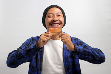 A happy young Asian man, dressed in a beanie hat and casual shirt, enthusiastically shows a credit card in his hand, feeling excited while demonstrating a solution for online shopping payment