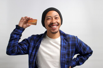A happy young Asian man, dressed in a beanie hat and casual shirt, enthusiastically shows a credit...