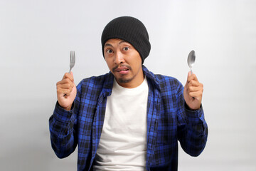 Hungry Asian man, dressed in a beanie hat and casual shirt, bites his lips while holding a fork and...