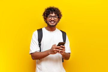 young indian guy student in glasses with backpack using smartphone on yellow isolated background
